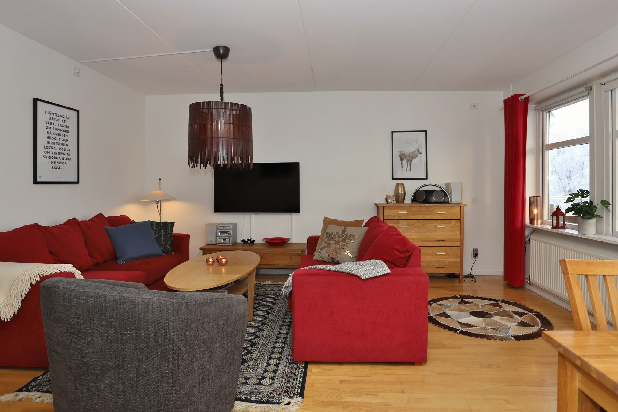 En hyggelig stue med røde sofaer, en grå lænestol, et sofabord i træ, et vægmonteret tv, indrammet kunst, en kommode i træ og et stort vindue med røde gardiner. Gulvet er af træ med tæpper.