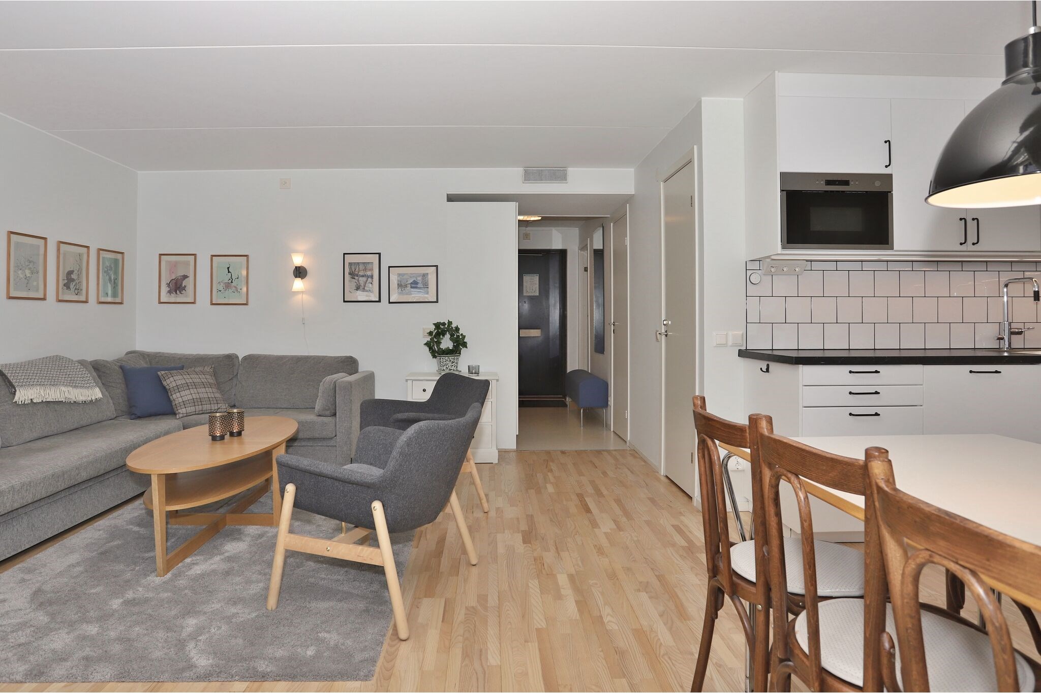 Moderne åben stue og køkken med lyse trægulve, grå sofa, lænestol, rundt sofabord, spisebord med træstole og hvide skabe. Vægkunst og stueplanter er en ekstra dekoration.