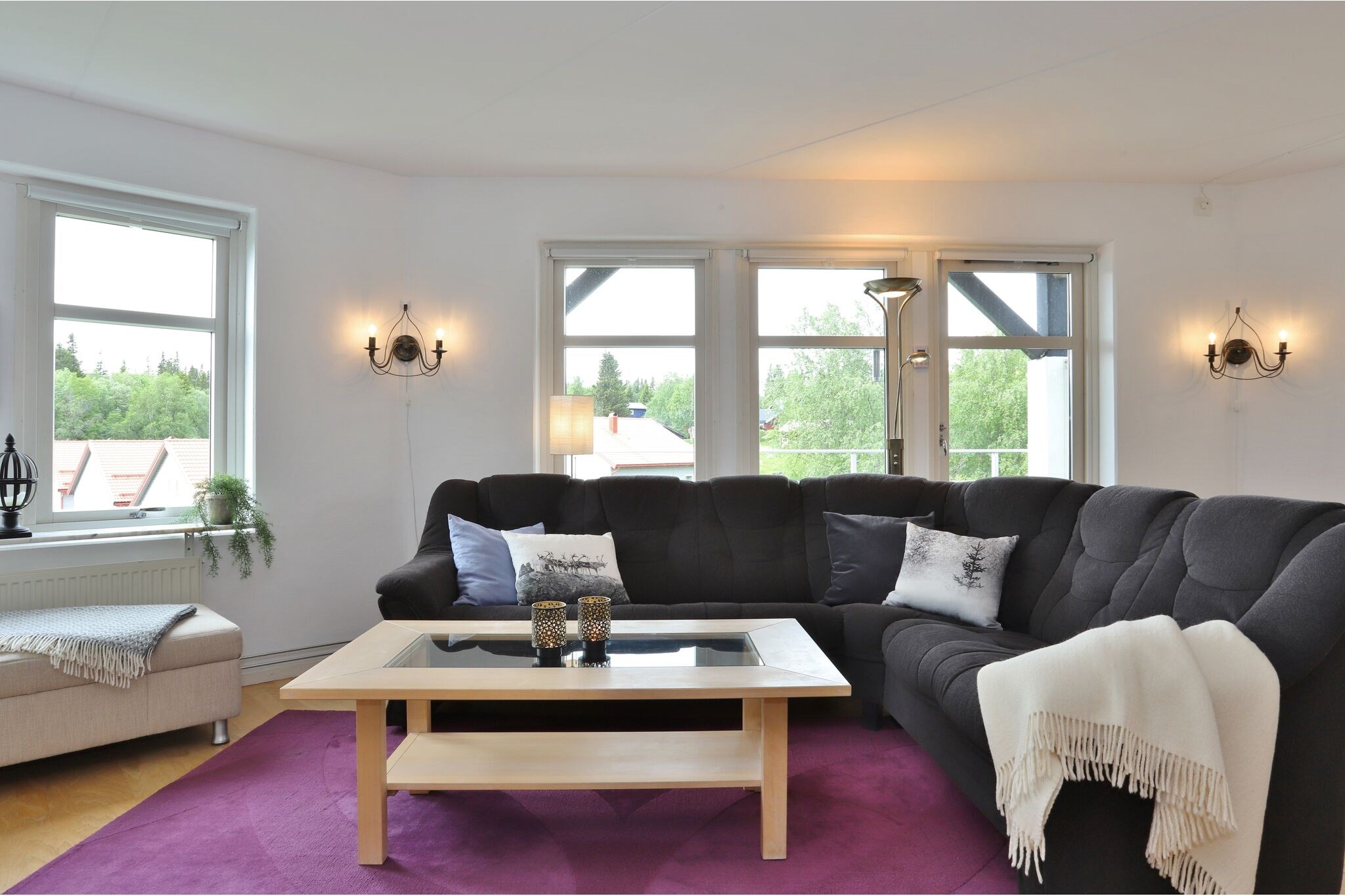 En hyggelig stue med en mørkegrå sofa, lyse tæpper, dekorative puder, et sofabord i træ og store vinduer, der lukker naturligt lys ind. Et lilla tæppe og en beige lænestol fuldender omgivelserne.
