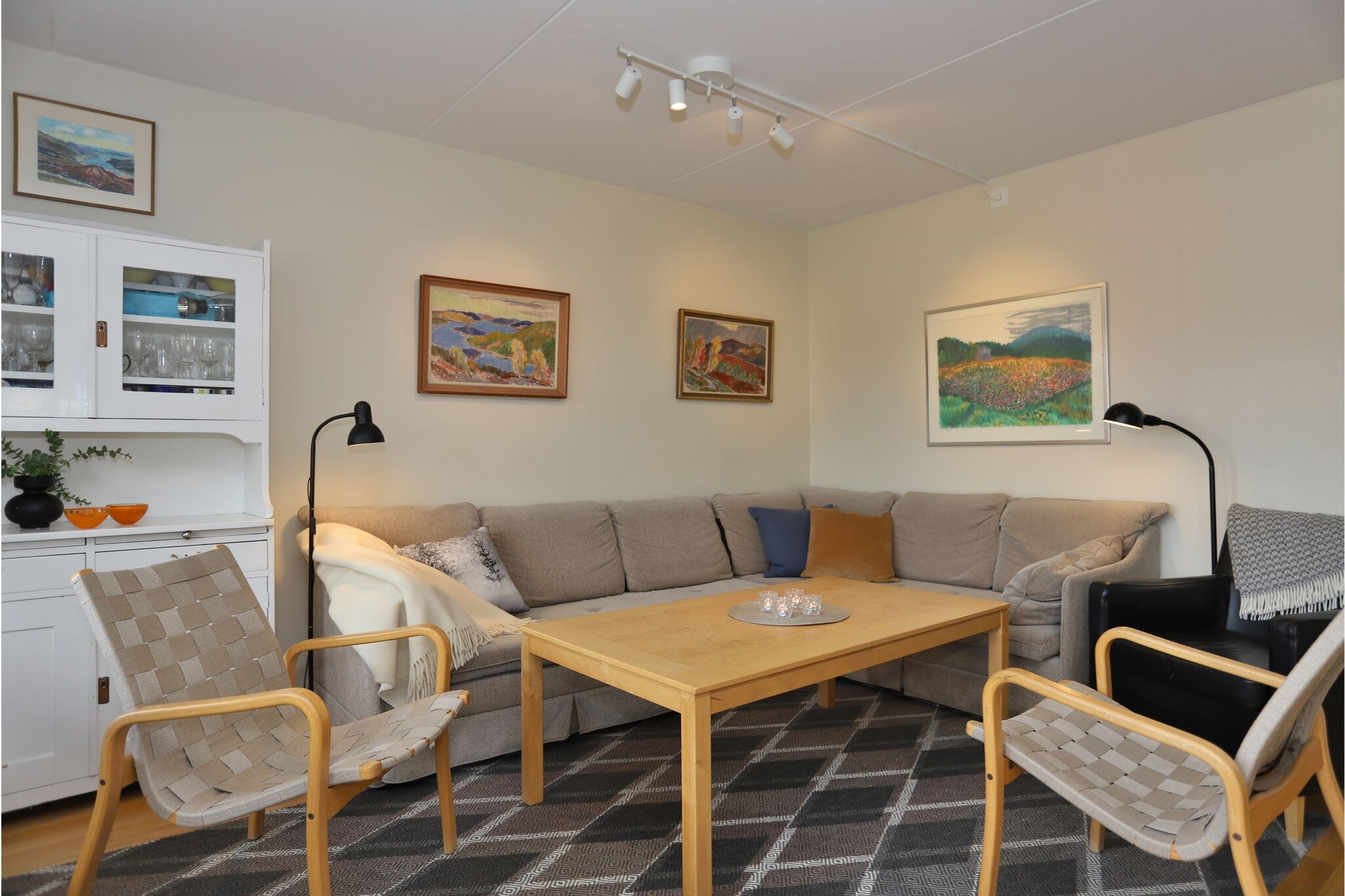 En hyggelig stue med en beige hjørnesofa, et sofabord i træ, flettede lænestole, en sort lænestol, vægkunst, et hvidt skabe og et gråt mønstret tæppe under bløde loftslamper.