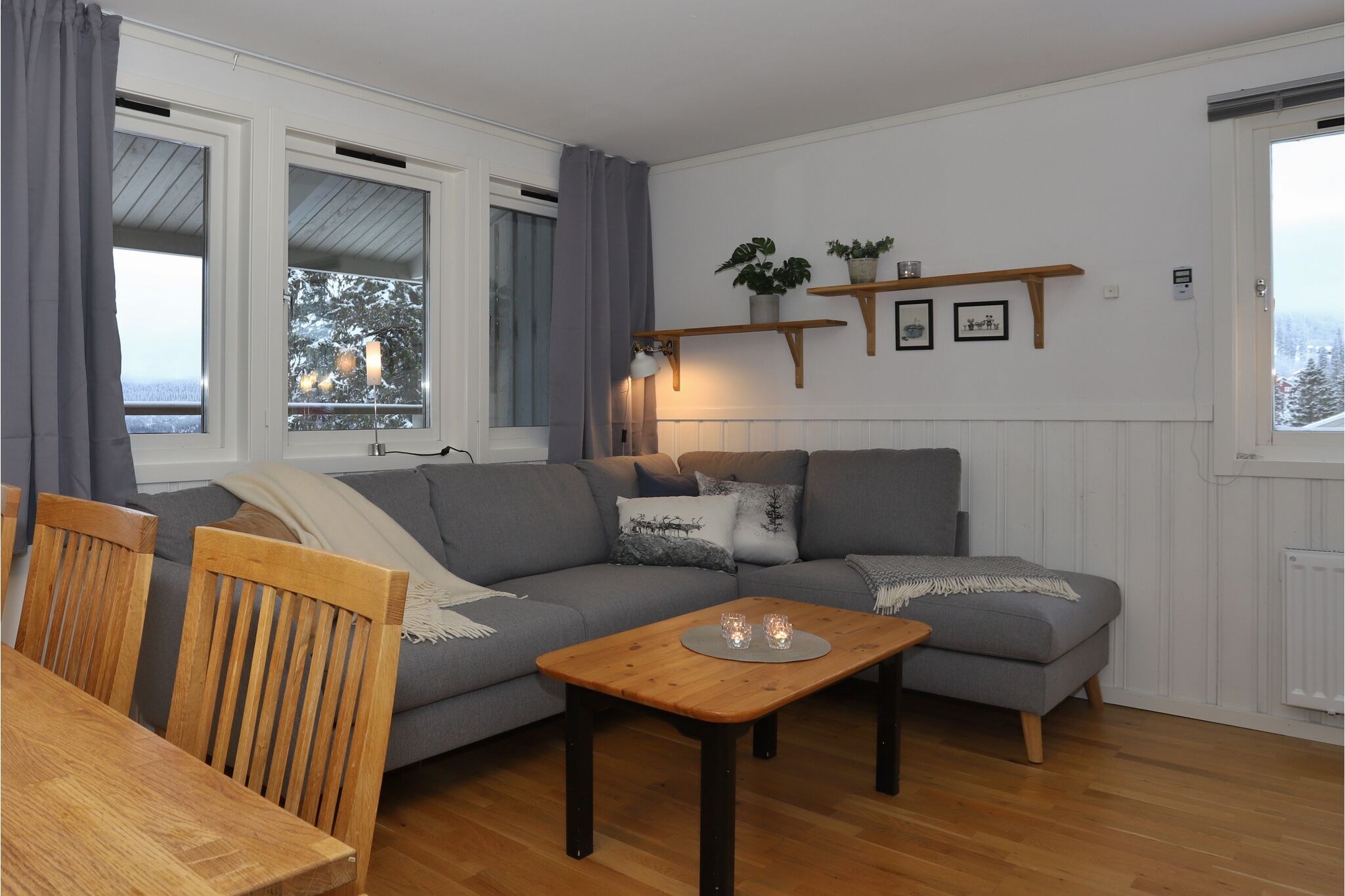 En hyggelig stue med en grå hjørnesofa, et sofabord i lyst træ og spisestuestole i træ. Grå gardiner, væghylder med planter og indrammet kunst pryder de hvide vægge. Store vinduer tilbyder udsigt til sneklædte træer udenfor.