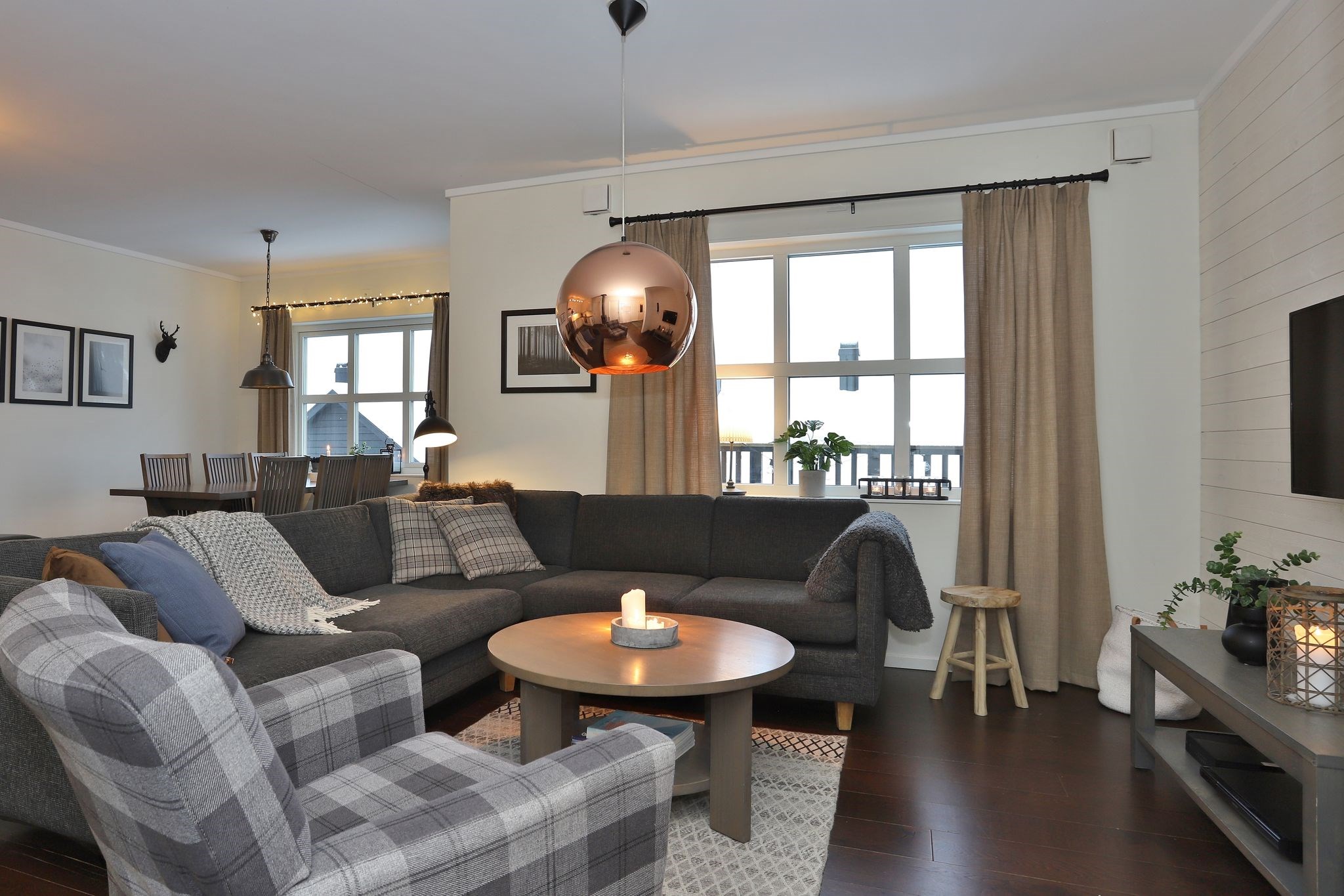 En hyggelig stue med en grå sektionssofa, en ternet lænestol, et rundt sofabord med et stearinlys, TV på væggen, store vinduer med taupefarvede gardiner og en spiseplads i baggrunden. Moderne og varm indretning overalt.