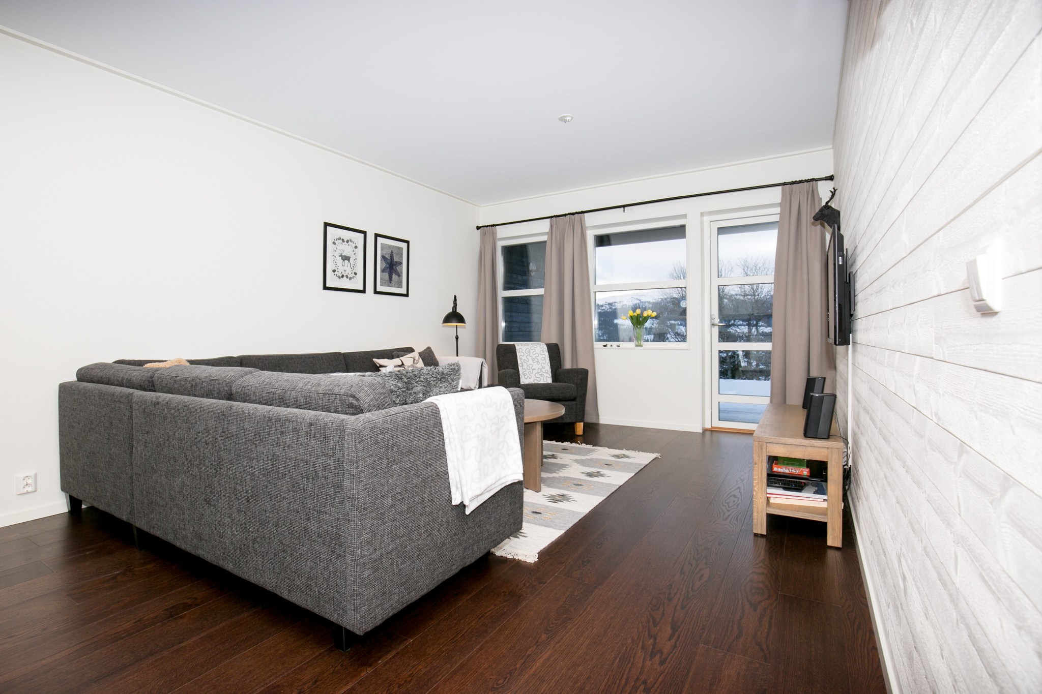 Moderne stue med mørke trægulve, en grå sofa, et lille bord, vægmonteret TV, et par indrammede billeder og store vinduer med beige gardiner med udsigt over et snedækket udendørs landskab.