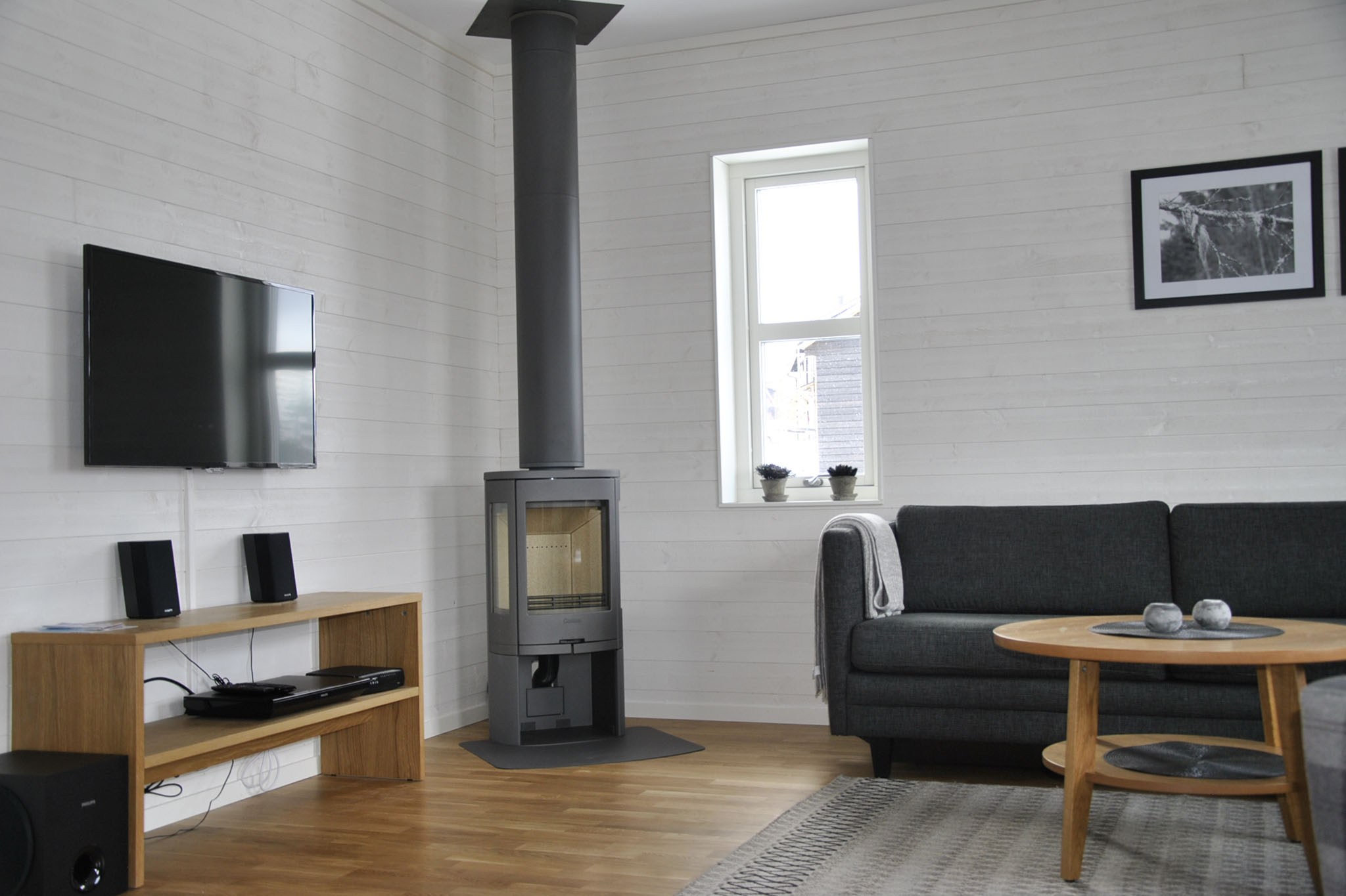 En moderne stue med hvide trævægge, en sort sofa, et rundt sofabord i træ, et vægmonteret tv, en brændeovn og et lille vindue, der lukker naturligt lys ind.