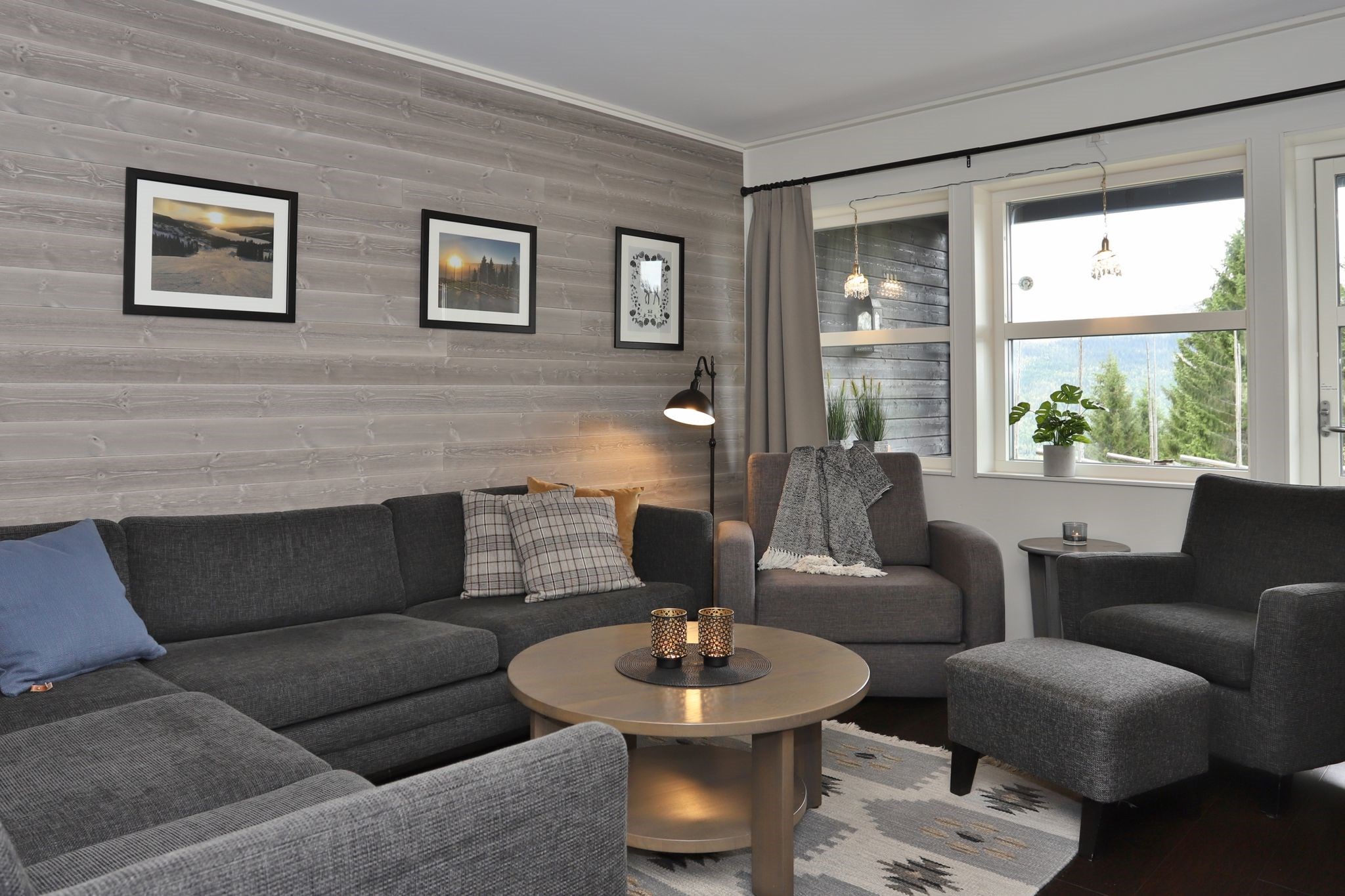En moderne stue med grå hjørnesofa, lænestol, rundt sofabord i træ med stearinlys, mønstret tæppe, indrammet kunst på en træpanelvæg og store vinduer med en smuk udsigt.