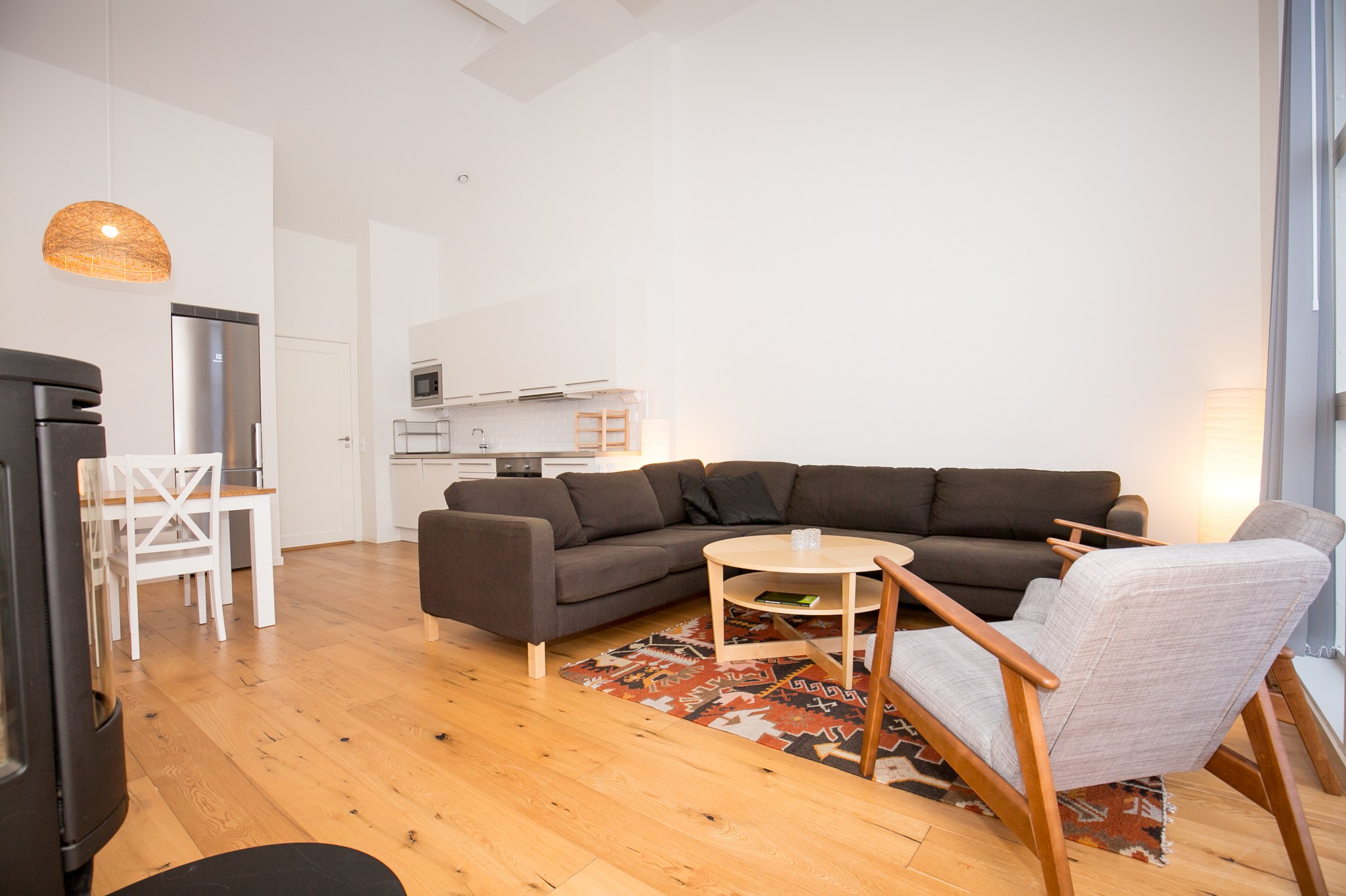 Moderne, lys stue med en stor mørk sofa, lys lænestol, rundt sofabord i træ, mønstret tæppe og åbent køkkenområde med hvide skabe og spisebord i baggrunden.