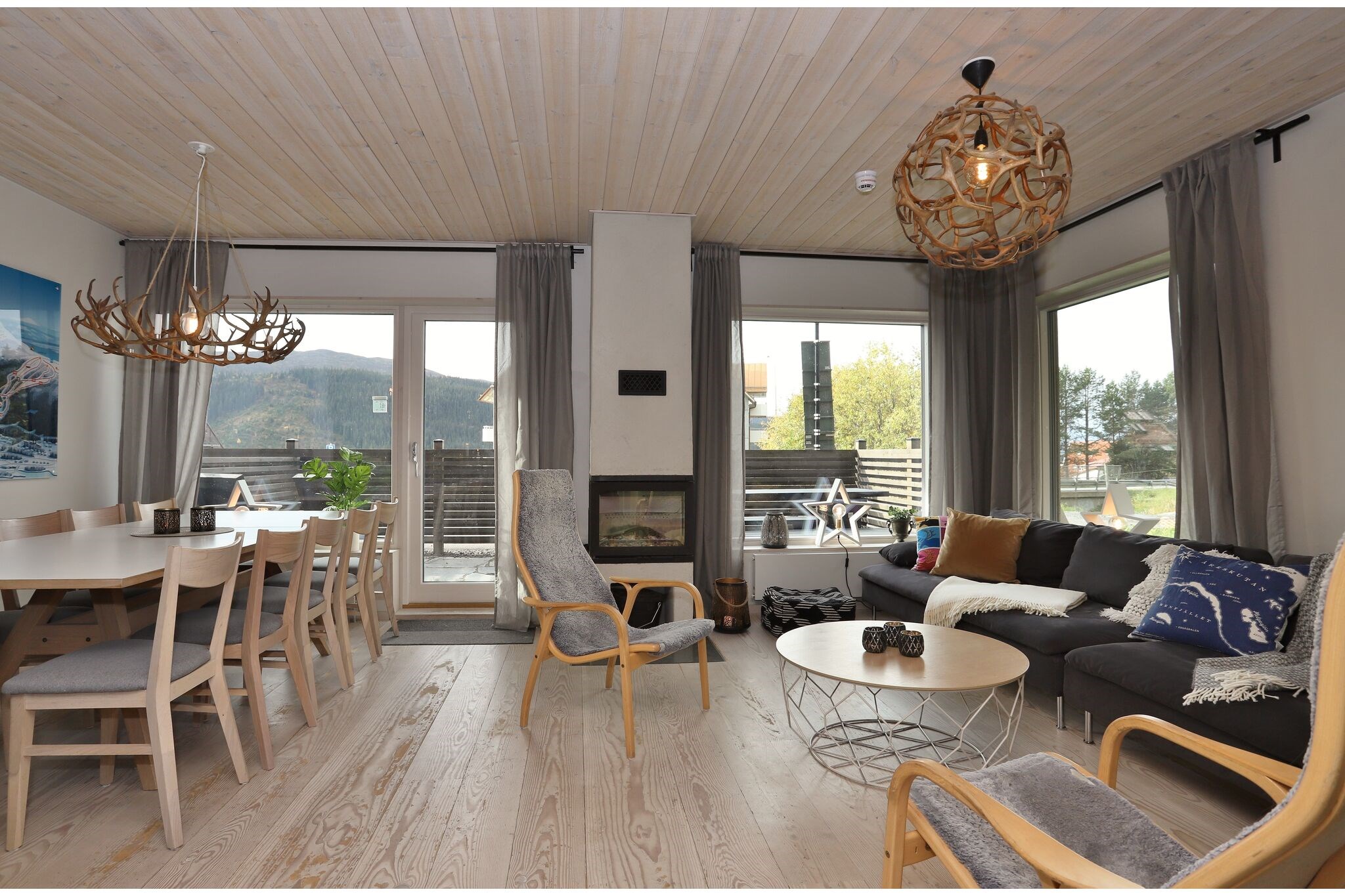 En moderne opholds- og spisestue med lyse trægulve og -lofter, store vinduer, grå gardiner, pejs, grå sofaer, træstole og to unikke pendellamper med udsigt over et bjerglandskab.