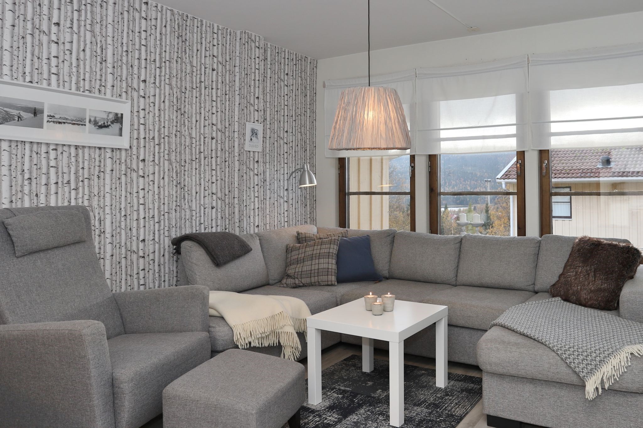 En hyggelig stue med grå sofaer, en grå lænestol, et hvidt sofabord, stearinlys og en gulvlampe. Væggen har et birketræsmønster, og store vinduer lukker naturligt lys ind med hvide persienner.