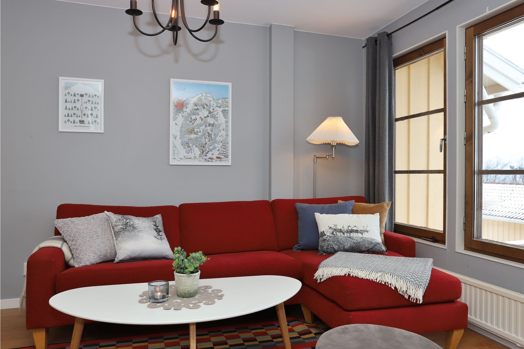 En moderne stue med en rød hjørnesofa, forskellige puder, et hvidt sofabord med planter og stearinlys, en gulvlampe, to indrammede billeder på en grå væg og store vinduer med naturligt lys.