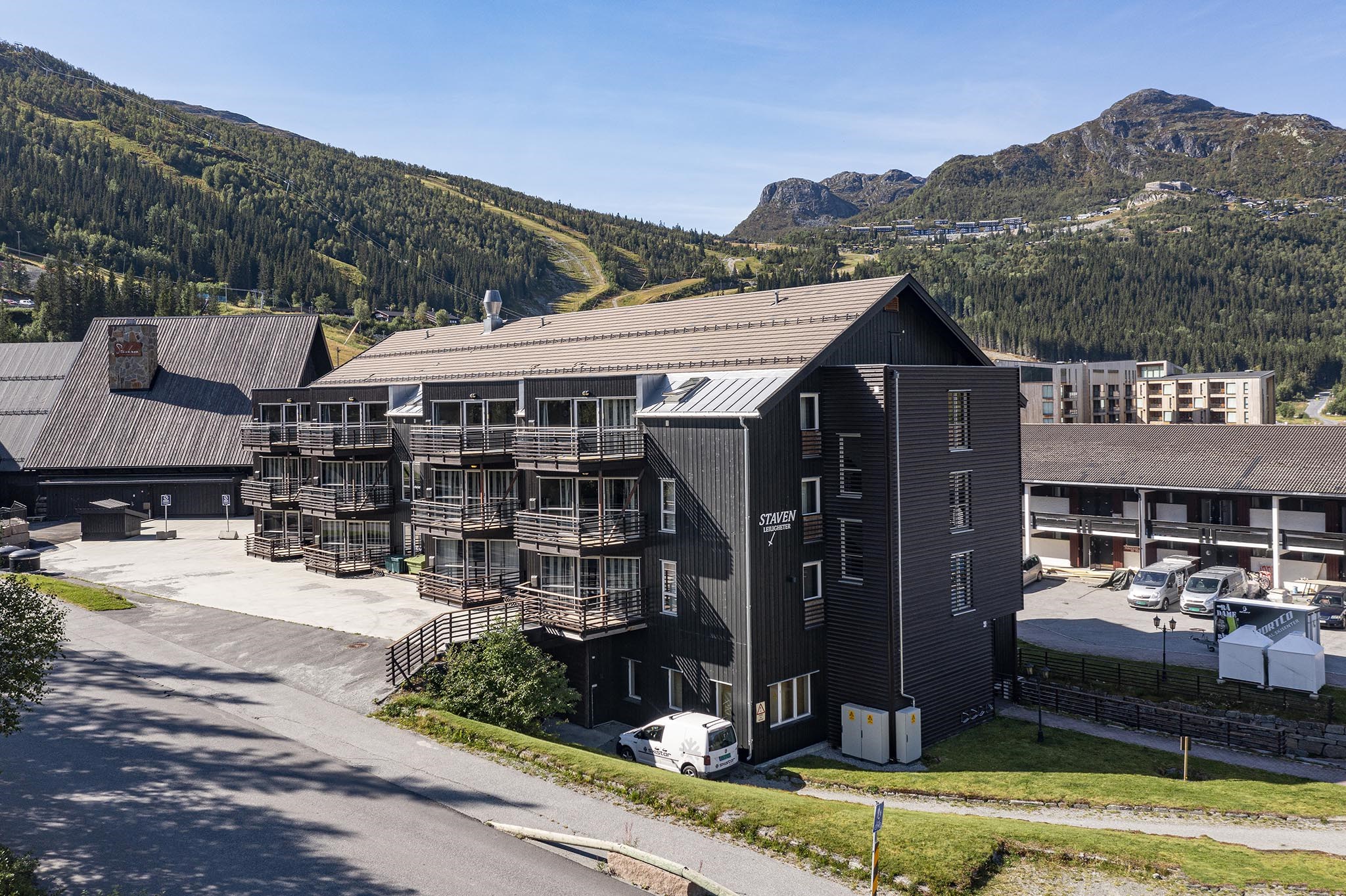 En moderne lejlighedsbygning i mørkt træ med altaner, omgivet af bjerge og grønne områder under en klar blå himmel. Adskillige parkerede biler og nærliggende bygninger er synlige i baggrunden.