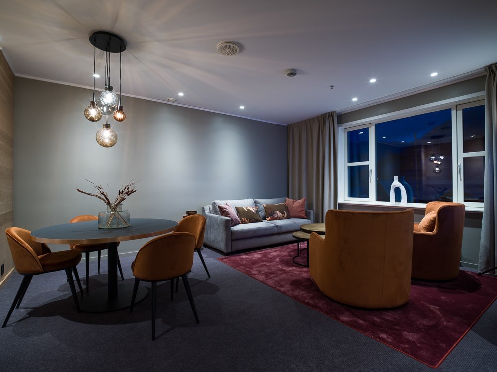 Moderne stue med en grå sofa, sennepsgule lænestole, et rundt spisebord med matchende stole, hængende pendellamper og et stort vindue med gardiner, alt sammen placeret på et mørkerødt tæppe.