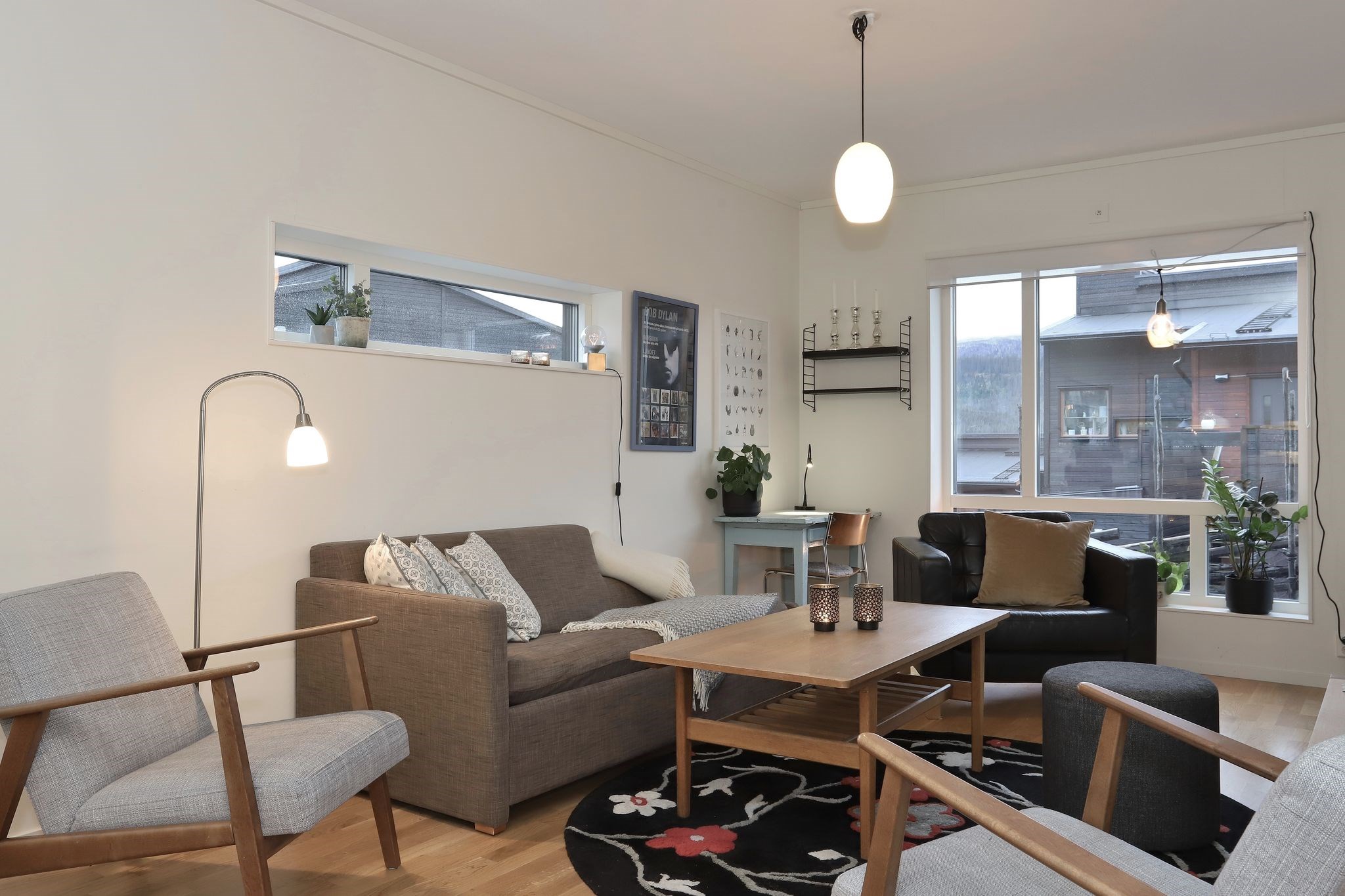 En moderne stue med en grå sofa, to lysegrå lænestole, en sort lænestol, et sofabord i træ og et mønstret tæppe. Store vinduer bringer naturligt lys ind, og planter og vægkunst pryder rummet.