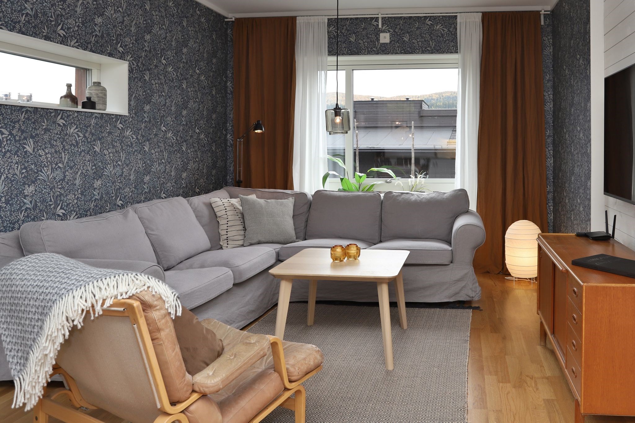 En hyggelig stue med en grå hjørnesofa, en beige lænestol, et sofabord i træ, mønstret tapet, brune gardiner, et tv på en træstander og en lysende gulvlampe ved vinduet.
