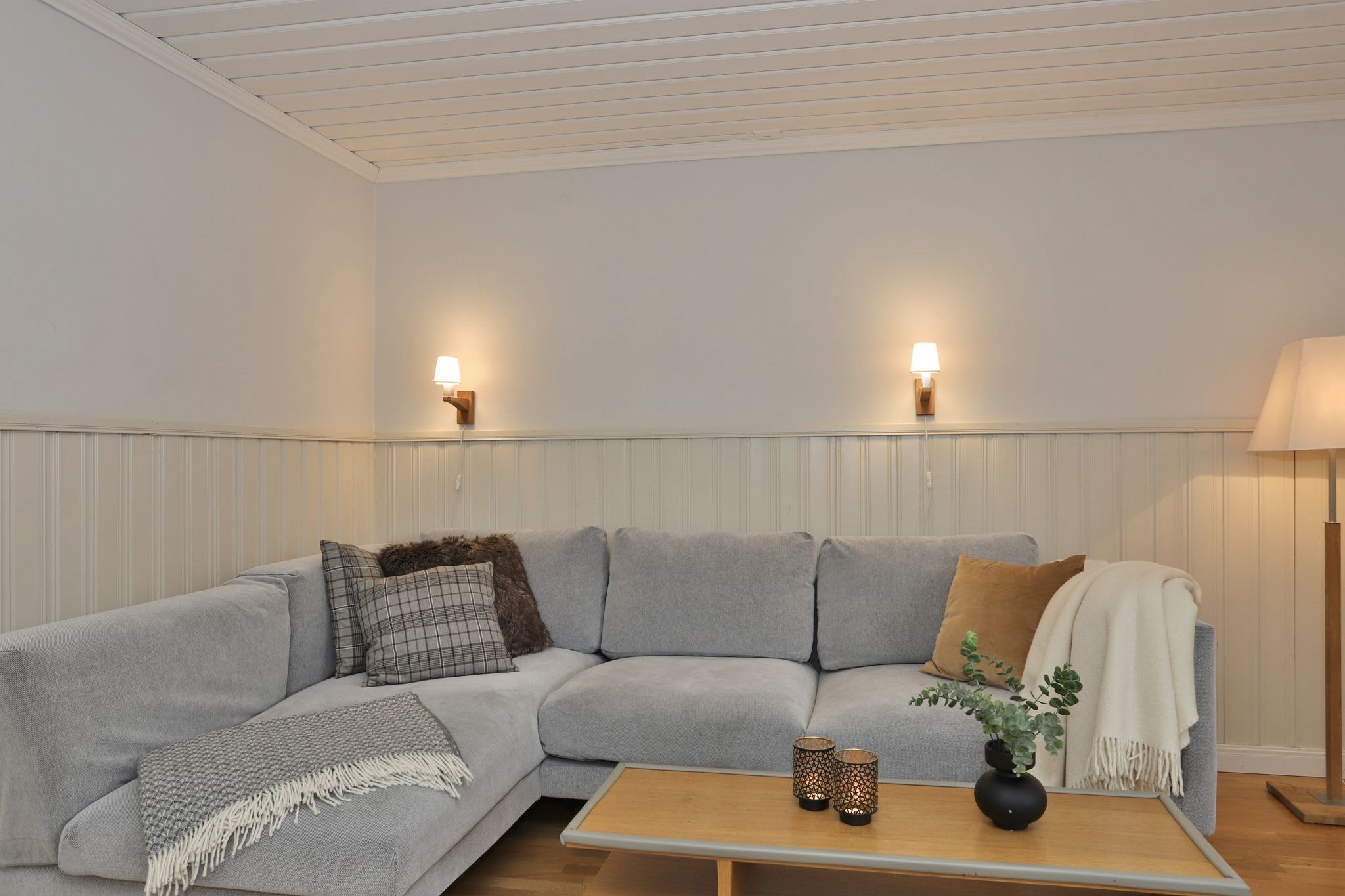 En hyggelig stue med en lysegrå sofa, ternede og teksturerede hynder, et tæppe, et sofabord i træ med stearinlys og en vase samt varme væg- og gulvlamper, der skaber en blød belysning.