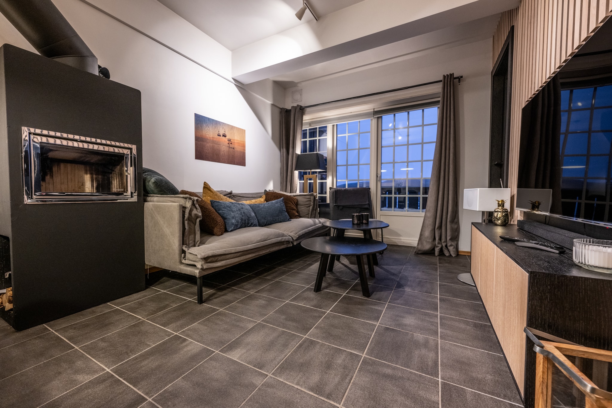 Moderne stue med gråt flisebelagt gulv, neutral sofa med blå og brune hynder, sorte borde, en pejs, store vinduer, vægkunst og minimalistisk indretning i blød belysning.