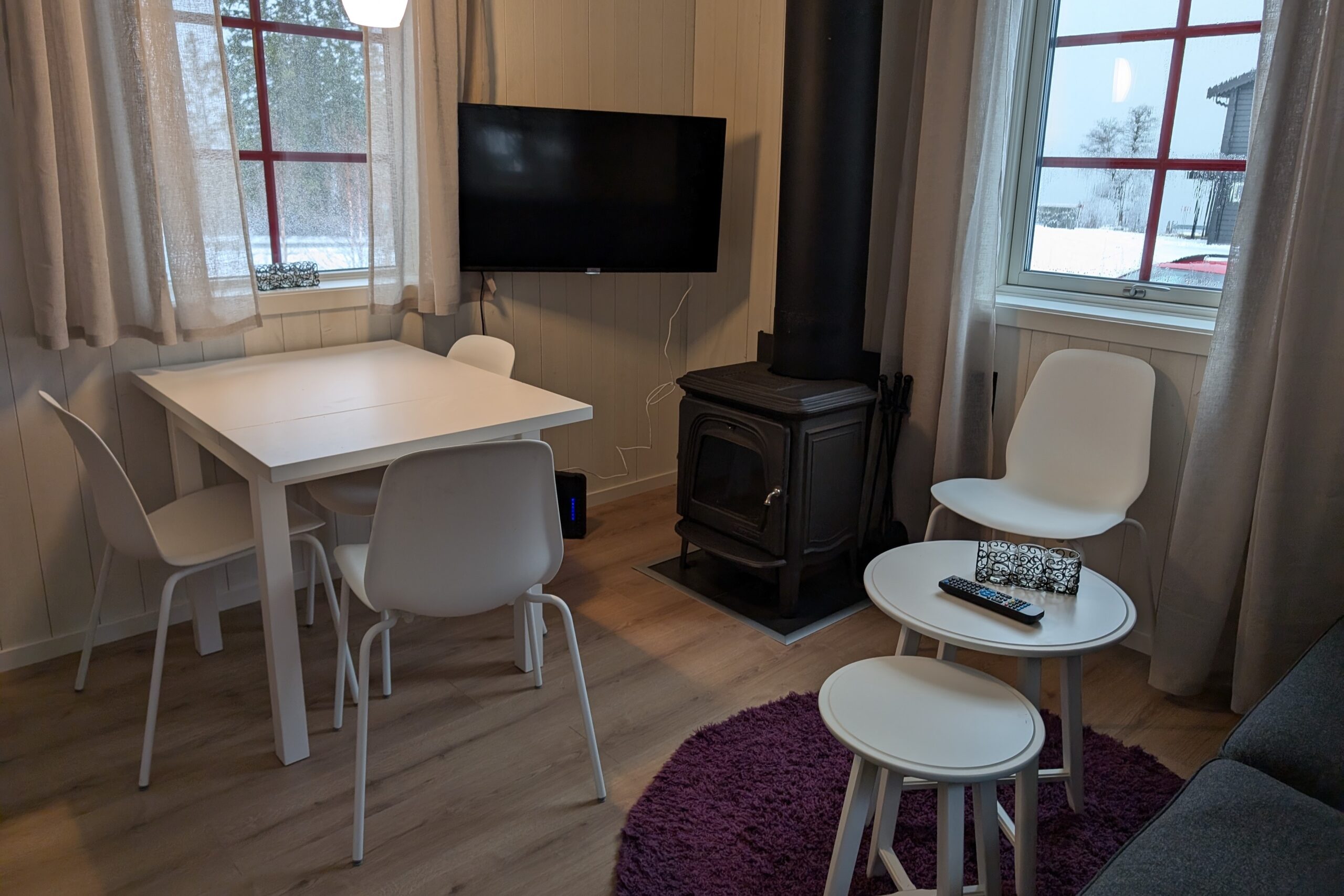 Et hyggeligt værelse med en brændeovn, et hvidt spisebord med fire stole, et vægmonteret tv, to runde sideborde og et vindue med et snedækket landskab udenfor. Lyse trægulve og lyse gardiner.
