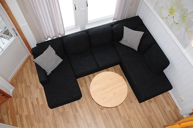 En stue set ovenfra og ned med en sort L-formet sofa, to sort-hvide stribede puder, et rundt sofabord i træ og lyst trægulv. Hvide gardiner og en blomstret vægkunst er også synlige.