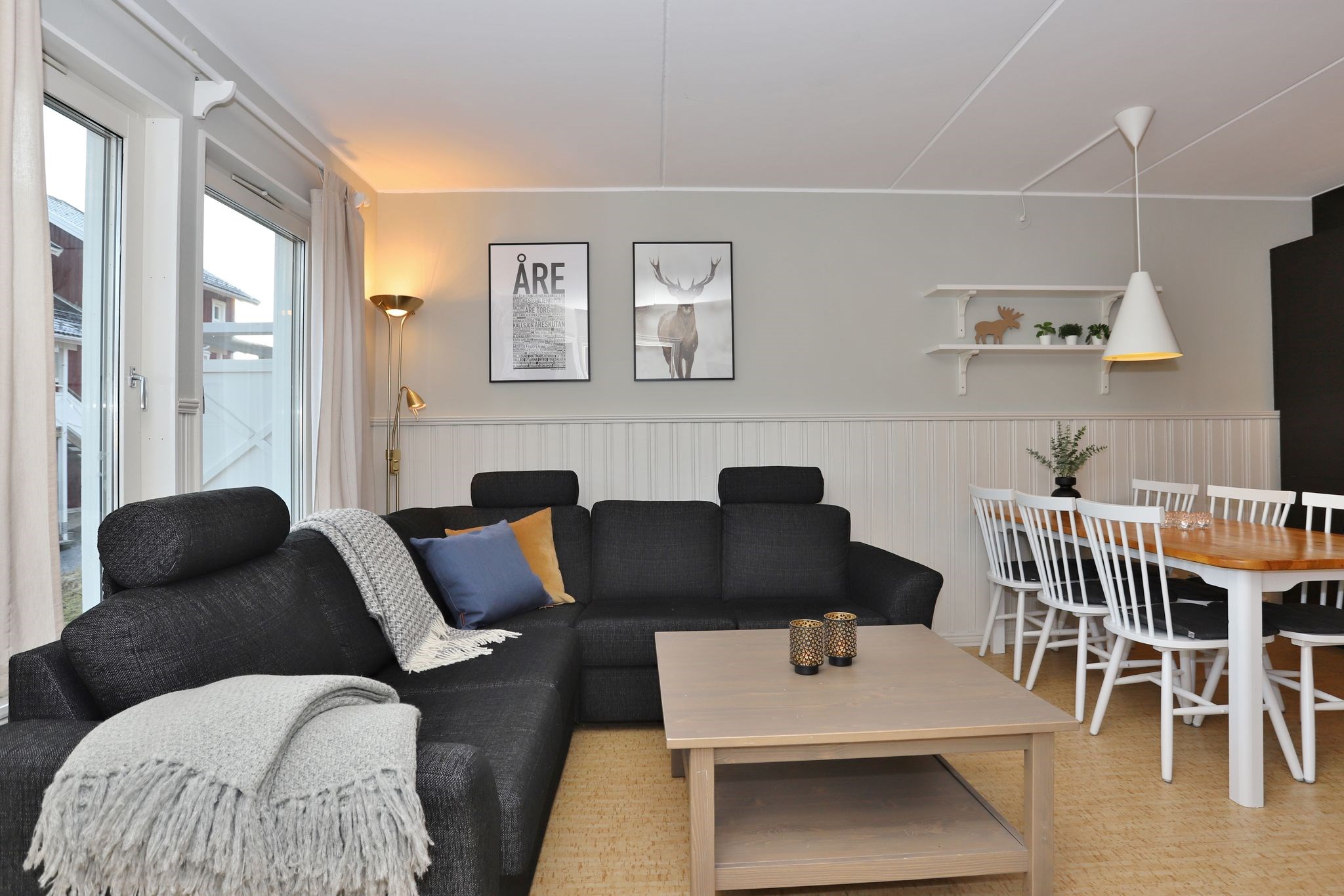 En moderne stue og spisestue med en sort sofa, sofabord i træ, indrammet vægkunst, hvide spisestuestole omkring et træbord, pendellampe og store vinduer, der lukker naturligt lys ind.