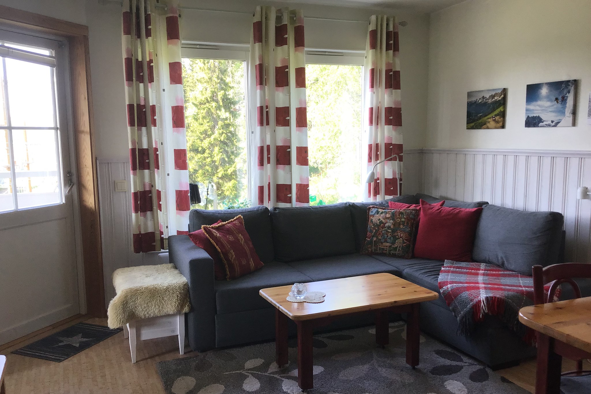 En hyggelig stue med en grå sofa, røde og mønstrede hynder, et rødt ternet tæppe, et sofabord i træ, rød- og hvidstribede gardiner, naturfotos på væggen og sollys, der strømmer ind gennem store vinduer.