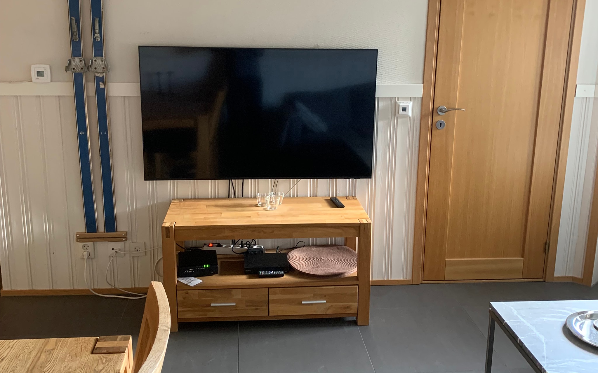Et fladskærms-tv er monteret på en væg over en trækonsol med elektronik, fjernbetjening og et glaspanel ovenpå. Der er synlige blå forsyningsrør til venstre og en lukket trædør til højre.