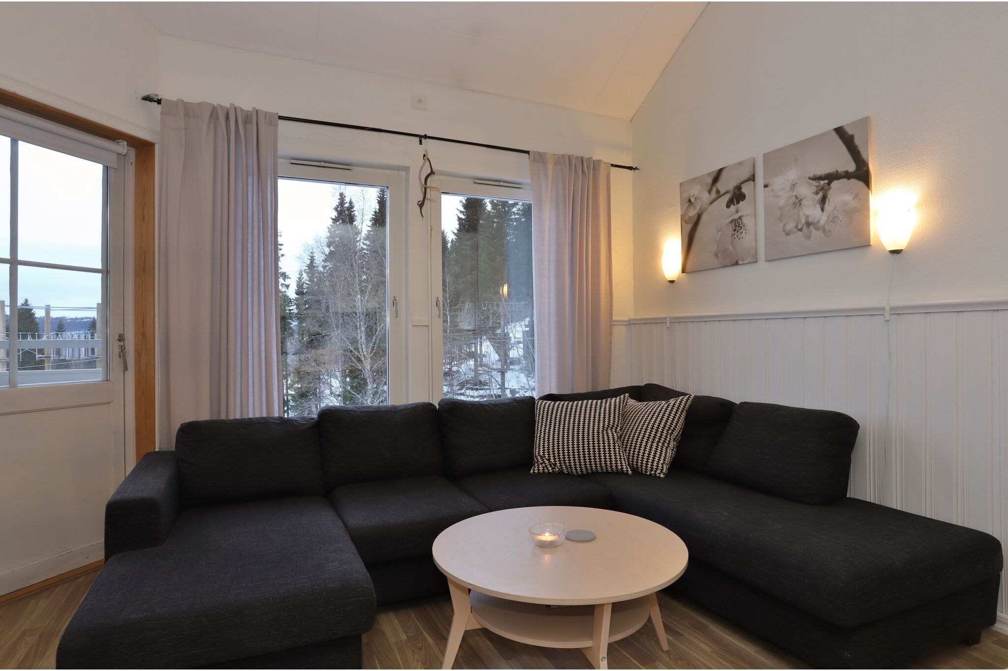 En hyggelig stue med en sort hjørnesofa, et rundt sofabord i lyst træ og et stearinlys. Store vinduer med lyse gardiner lukker naturligt lys ind og viser sneklædte træer udenfor. To blomsterkunstværker hænger på en lys væg.