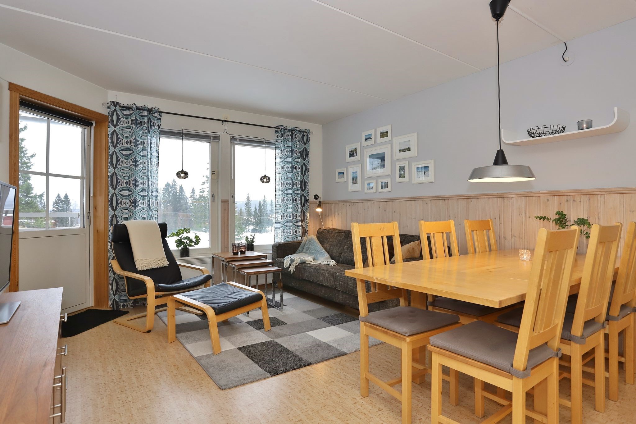 En hyggelig opholds- og spisestue med træmøbler, en grå sofa, en mønstret lænestol, et rektangulært spisebord med otte stole, store vinduer med blå gardiner og indrammede fotos på en lys væg.