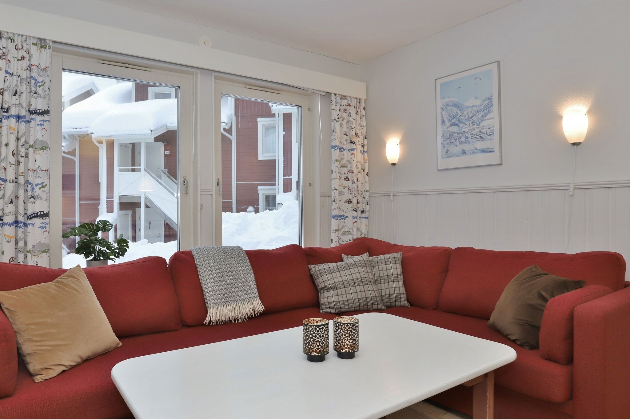 En hyggelig stue med en rød hjørnesofa, forskellige hynder, et hvidt sofabord med stearinlys, mønstrede gardiner, væglamper og et snedækket landskab, der kan ses gennem de store vinduer.