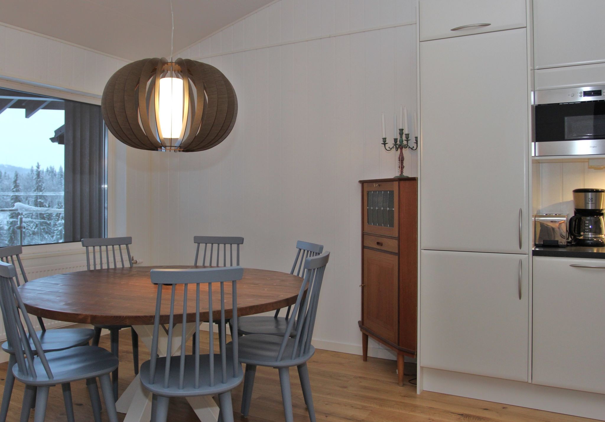 En moderne spiseplads med et rundt træbord omgivet af seks grå stole, en stor pendellampe ovenover, et vindue med udsigt over sneen, et lille træskab og hvide køkkenskabe med indbyggede apparater.
