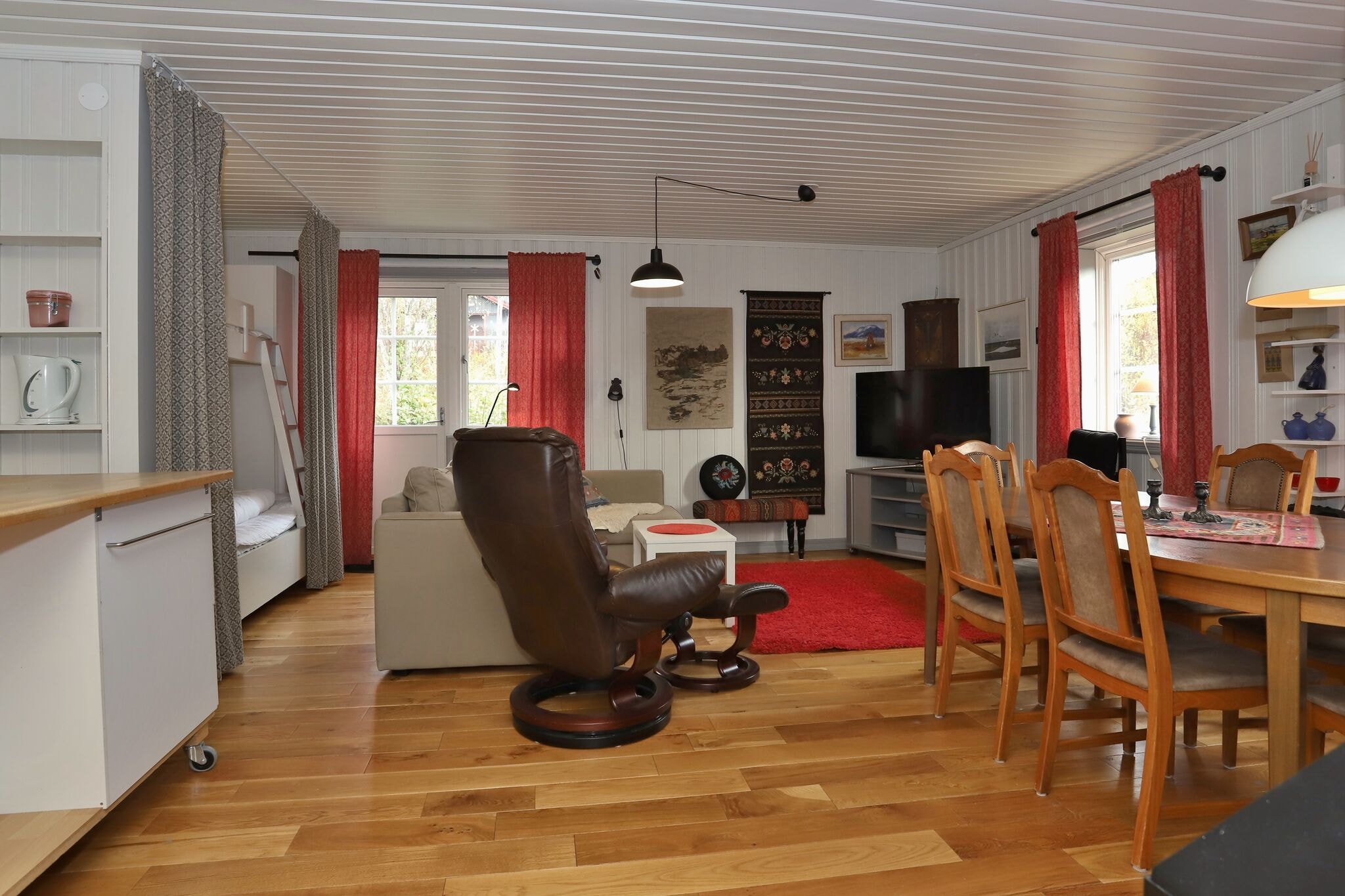 En hyggelig opholds- og spisestue med trægulve, en beige sofa, brun lænestol, spisebord i træ med stole, tv, bogreoler og røde gardiner for vinduerne; varm og indbydende indretning med vægkunst og et rødt tæppe.