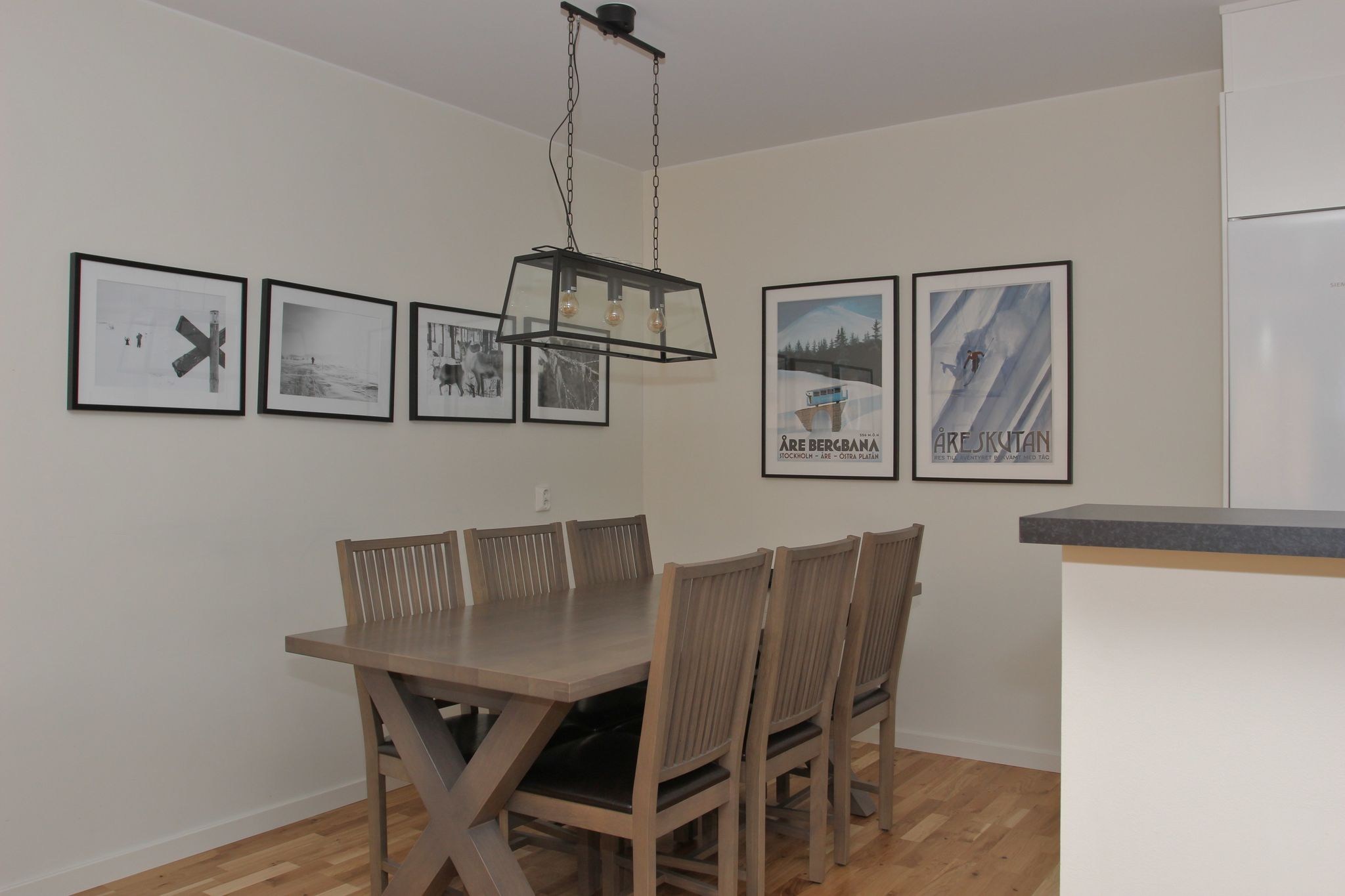 En moderne spiseplads med et træbord og otte matchende stole, vægkunst med sort-hvide og farvetryk, lyst trægulv og en rektangulær lampe med sort indrammet lofts.