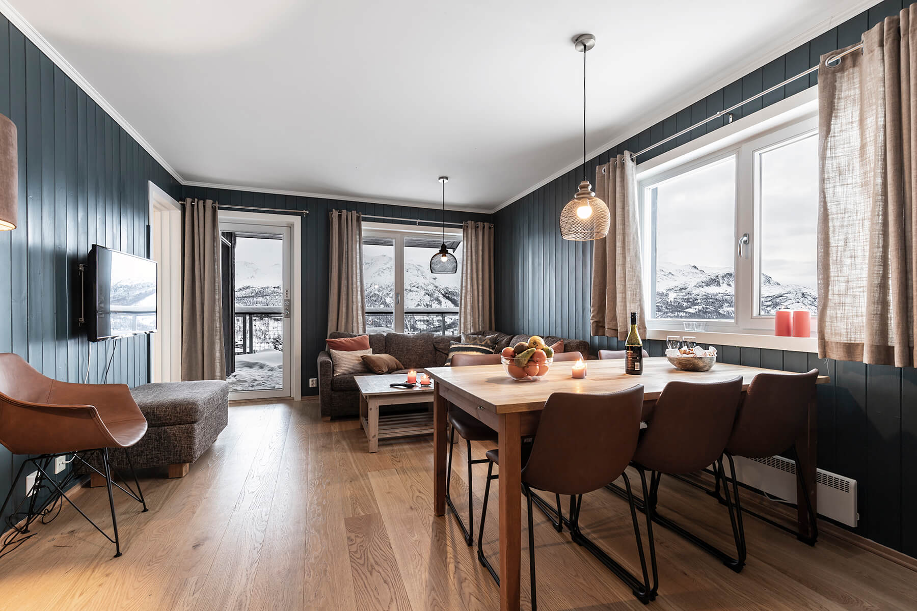 Moderne opholds- og spisestue med trægulve, mørke panelvægge, en hyggelig sofa, spisebord med fire stole og store vinduer med udsigt over sneklædte bjerge. Frugt og stearinlys pynter bordet.