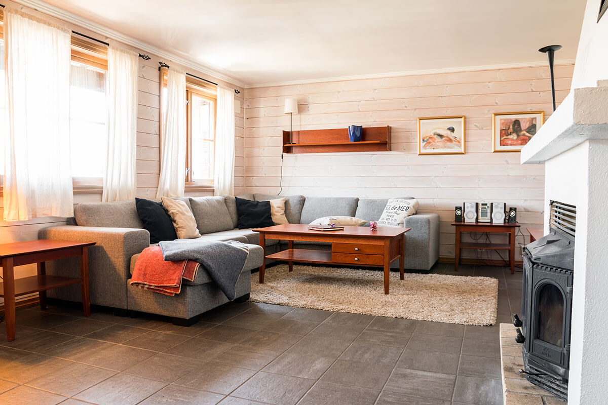 En hyggelig stue med en grå sofa, et sofabord i træ og et beige tæppe. Sollys strømmer ind gennem store vinduer. Kunst hænger på de hvide panelvægge, og en brændeovn står i hjørnet ved siden af et stereoanlæg.