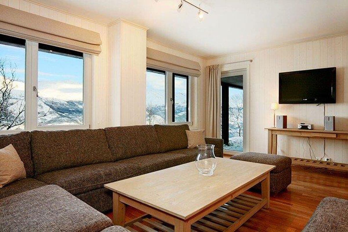 En hyggelig stue med store vinduer med udsigt over sneklædte bjerge, en grå hjørnesofa, et sofabord i træ med en glasvase, et vægmonteret tv og et konsolbord med højttalere og en lampe.