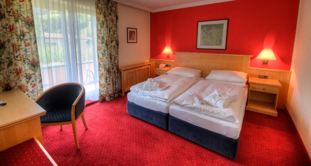 Et hyggeligt hotelværelse med to enkeltsenge skubbet sammen, rødt tæppe, rød accentvæg, væglamper, blomstrede gardiner, et skrivebord med en stol og et stort vindue, der fører ud til en balkon.