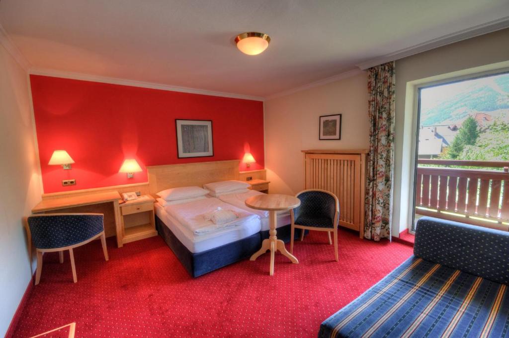 Et hyggeligt hotelværelse med en dobbeltseng, to stole, et lille rundt bord, et skrivebord og en sofa. Værelset har rødt gulvtæppe, en rød accentvæg, væglamper og en balkon med udsigt over grønne områder og bjerge.