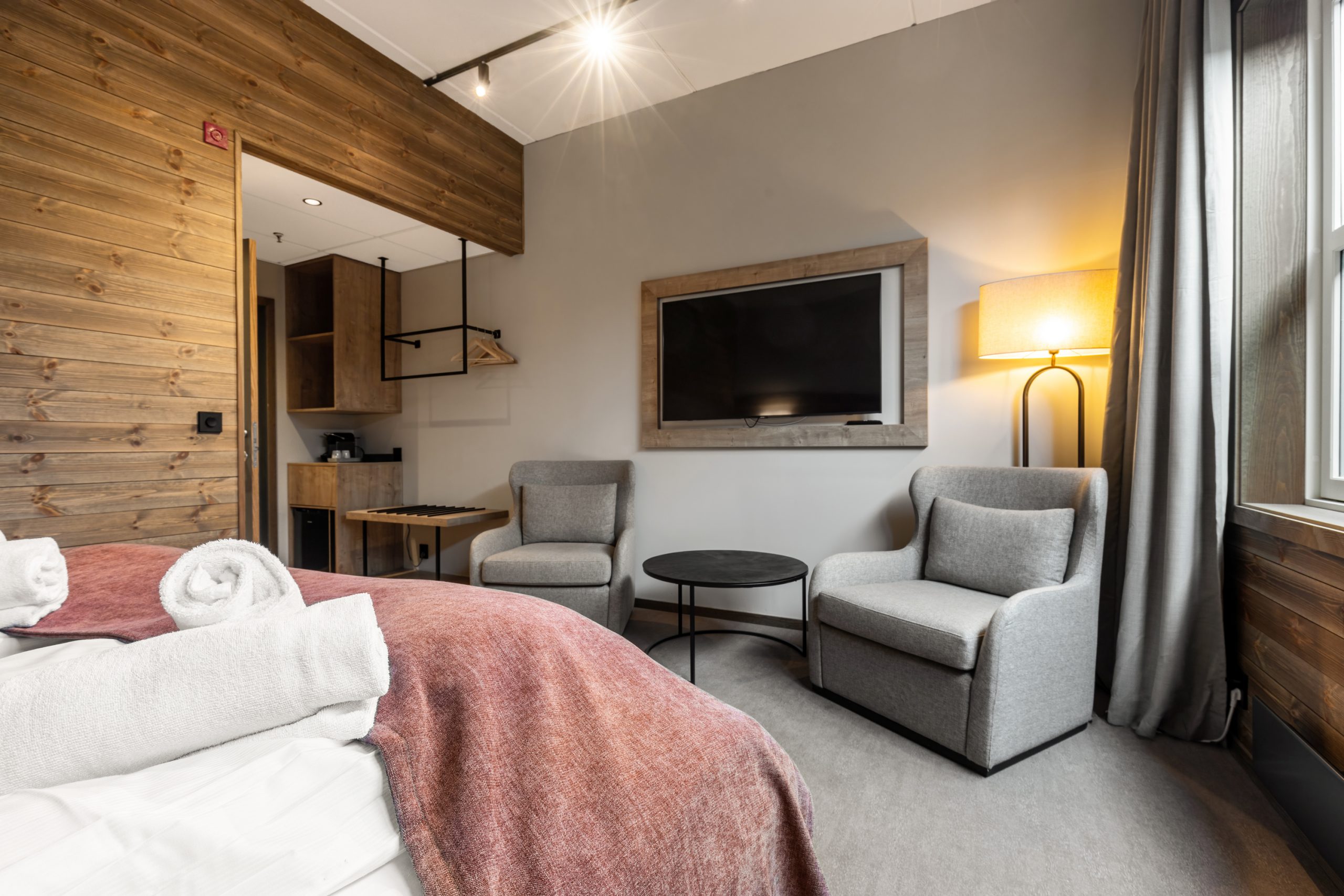 Moderne hotelværelse med to grå lænestole, et rundt sofabord, et stort vægmonteret tv, en høj gulvlampe og en seng med hvidt sengetøj og et rødligt tæppe. Trædetaljer og et vindue skaber en hyggelig atmosfære.