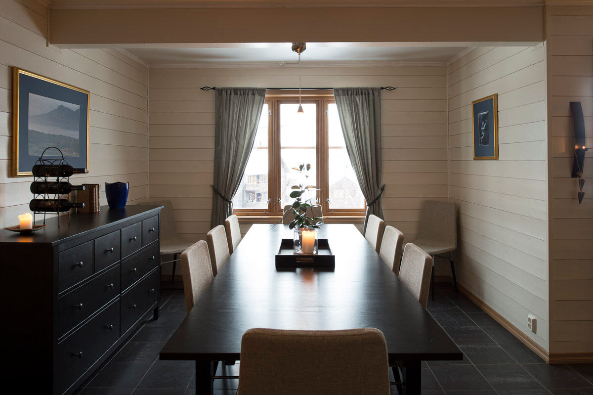 En moderne spisestue med et langt mørkt bord, otte stole, et midtpunkt med stearinlys og planter. Lyse træpaneler på væggene, indrammet kunst, en skænk med dekoration og et stort vindue med gardiner, der lukker naturligt lys ind.