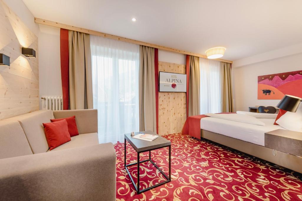Et lyst hotelværelse med en dobbeltseng, en beige sofa med røde hynder, et lille sofabord, et tæppe med rødt og guldmønstret mønster, et stort vindue med gardiner og moderne kunst på væggen.