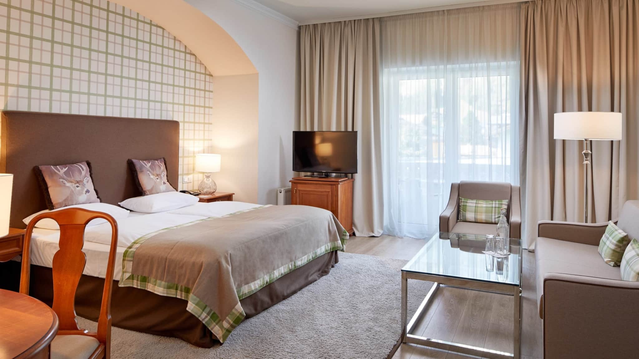 Et moderne hotelværelse med en dobbeltseng, to puder, sengelamper, et tv på et træbord, en lænestol, en sofa, et sofabord af glas med vandflasker, lyse gardiner og indretning i neutrale toner.