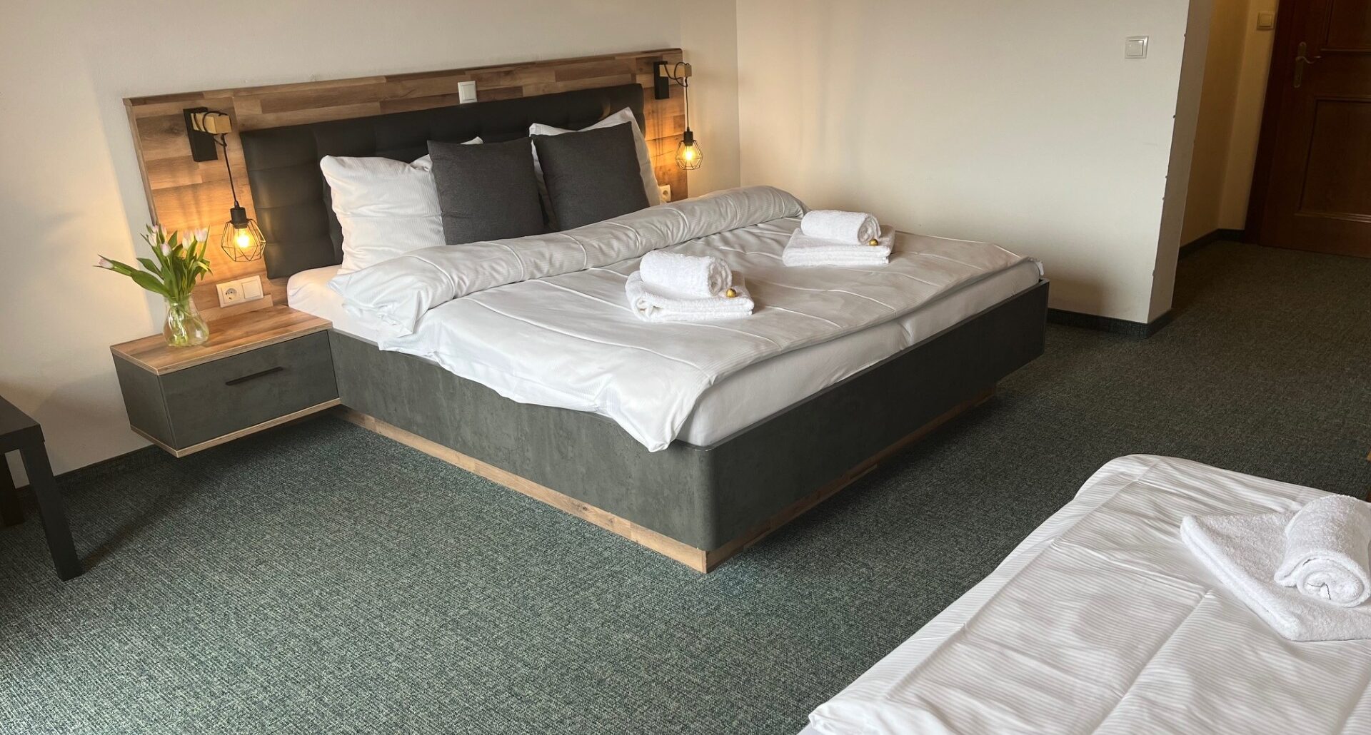 Et hotelværelse med en stor, pænt redt seng, to natborde med lamper, rene håndklæder placeret på sengen og en vase med blomster. En anden mindre seng er delvist synlig i forgrunden.