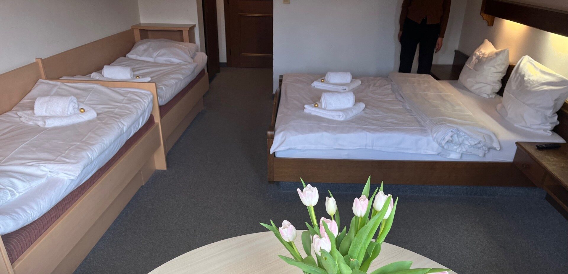 Et hotelværelse med to enkeltsenge og en dobbeltseng, alle pænt opredt med hvidt sengetøj og håndklæder. Et rundt bord med en vase med lyserøde tulipaner er i forgrunden. En person står nær bagvæggen.