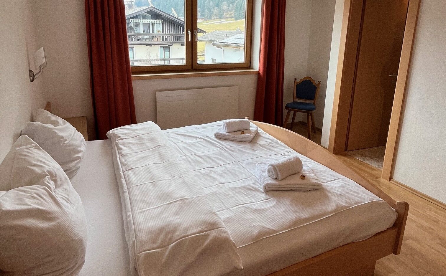 Et ryddeligt hotelværelse med en dobbeltseng, hvidt sengetøj, to foldede håndklæder, et vindue med røde gardiner, trægulve, en blå stol og en åben dør til et badeværelse.