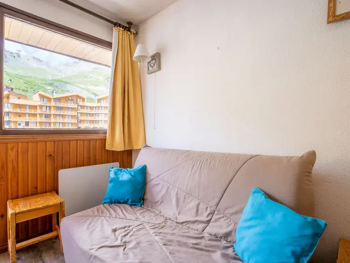 En beige sofa med to blå hynder står op ad en hvid væg ved siden af en lille træskammel. Et stort vindue med gule gardiner afslører udsigt over en bygning og bjerge udenfor.