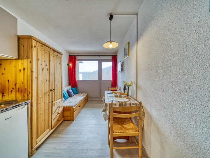 Lille, lyst atelier med træmøbler, en dagseng med blå hynder, spisebord med to stole og et stort vindue med røde gardiner, der lukker naturligt lys ind.
