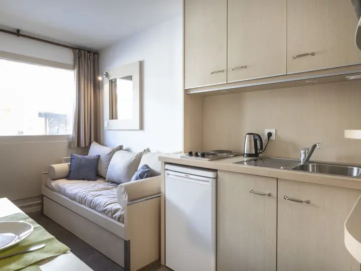 Moderne studiolejlighed med et kompakt tekøkken med vask, minikøleskab, komfur og elkedel. En beige sofa med hynder står ved et vindue med brune gardiner, der lukker naturligt lys ind.