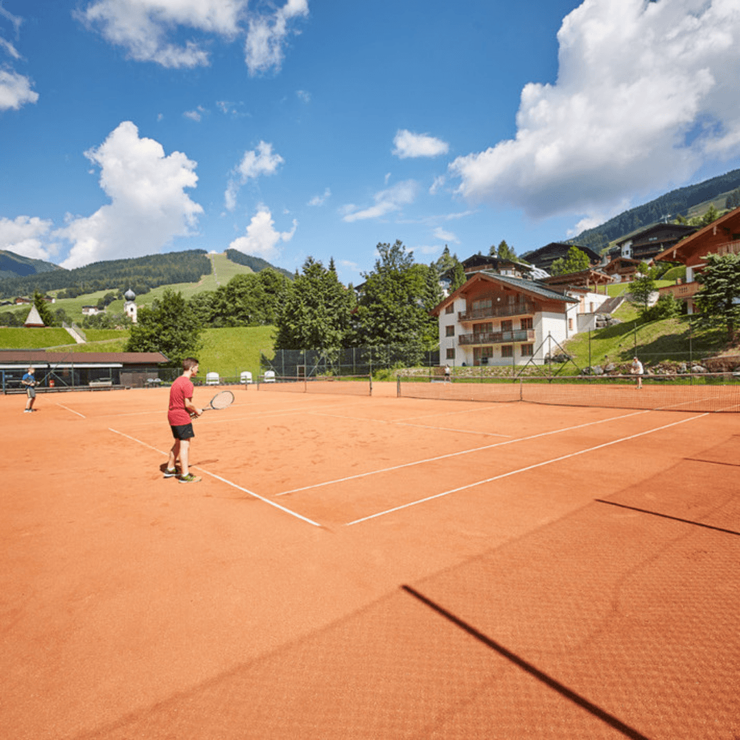 To personer, der spiller tennis på en lerbane i Westendorf, omgivet af frodige grønne bakker og bygninger i alpestil, under en klar blå himmel med spredte skyer; det er de perfekte rammer for en sommerferie i Østrig.