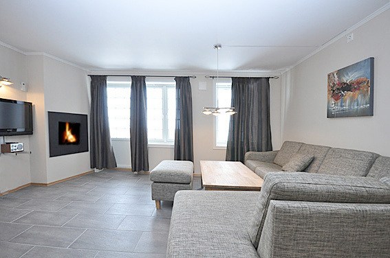 En moderne stue med en grå sektionssofa, matchende skammel og et sofabord i træ på et klinkegulv fremkalder den hyggelige charme ved en Westendorf skiferie. Værelset har et væghængt tv, pejs og to store vinduer med grå gardiner. Et maleri hænger på væggen.