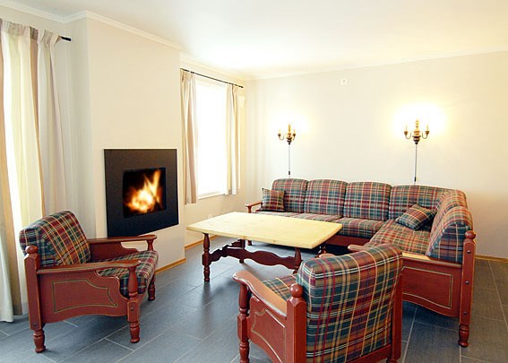 En hyggelig stue i stil med et charmerende Westendorf-retræte med plaidmønstrede sofaer og lænestol, et sofabord i træ og en pejs med flammer på venstre væg. To væglamper giver lys over siddeområdet. Cremefarvede gardiner pryder vinduerne.