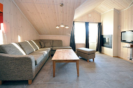 En hyggelig stue med skrå trælofter og vægge, der minder om en Westendorf skiferie. Den har en grå L-formet sofa, et sofabord i træ, en matchende skammel, et vægmonteret tv, en pejs og store vinduer prydet med sorte gardiner. Gulvet er elegant flisebelagt.