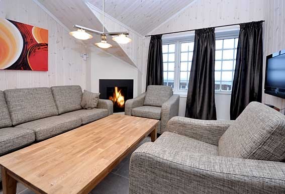 En hyggelig stue med lyse træpaneler, der minder om et skiferiested i Saalbach, har en lysegrå sofa og lænestole omkring et sofabord i træ. En pejs er tændt til venstre, med et moderne kunstmaleri ovenfor. Mørke gardiner indrammer et vindue til højre.